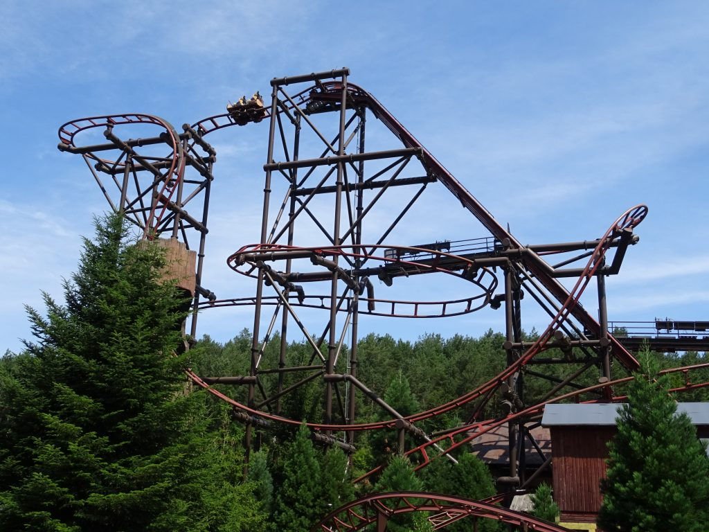 Sur la photo de Timber Drop, on distingue une imposante structure métallique en forme de tronc d'arbre, culminant à 30 mètres de hauteur. Au sommet, un train de montagnes russes est suspendu, prêt à entamer une descente vertigineuse.